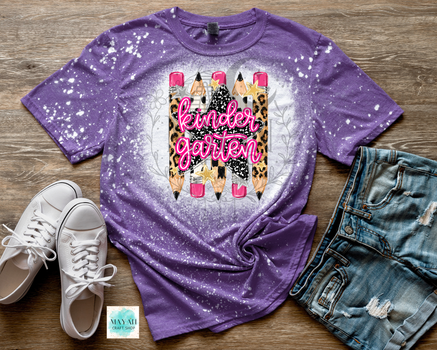 A purple short-sleeve t-shirt with a white splatter bleach effect and a central graphic of pencils with leopard print, pink lettering reading 'kindergarten', and a white circular glow. The shirt is styled in a flat lay on a wood background with white sneakers, denim shorts.