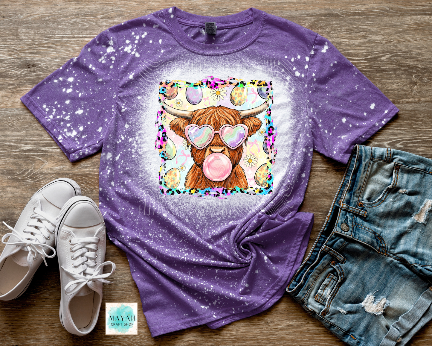 Purple t-shirt with a cartoon cow design on a wooden surface with shoes and shorts.