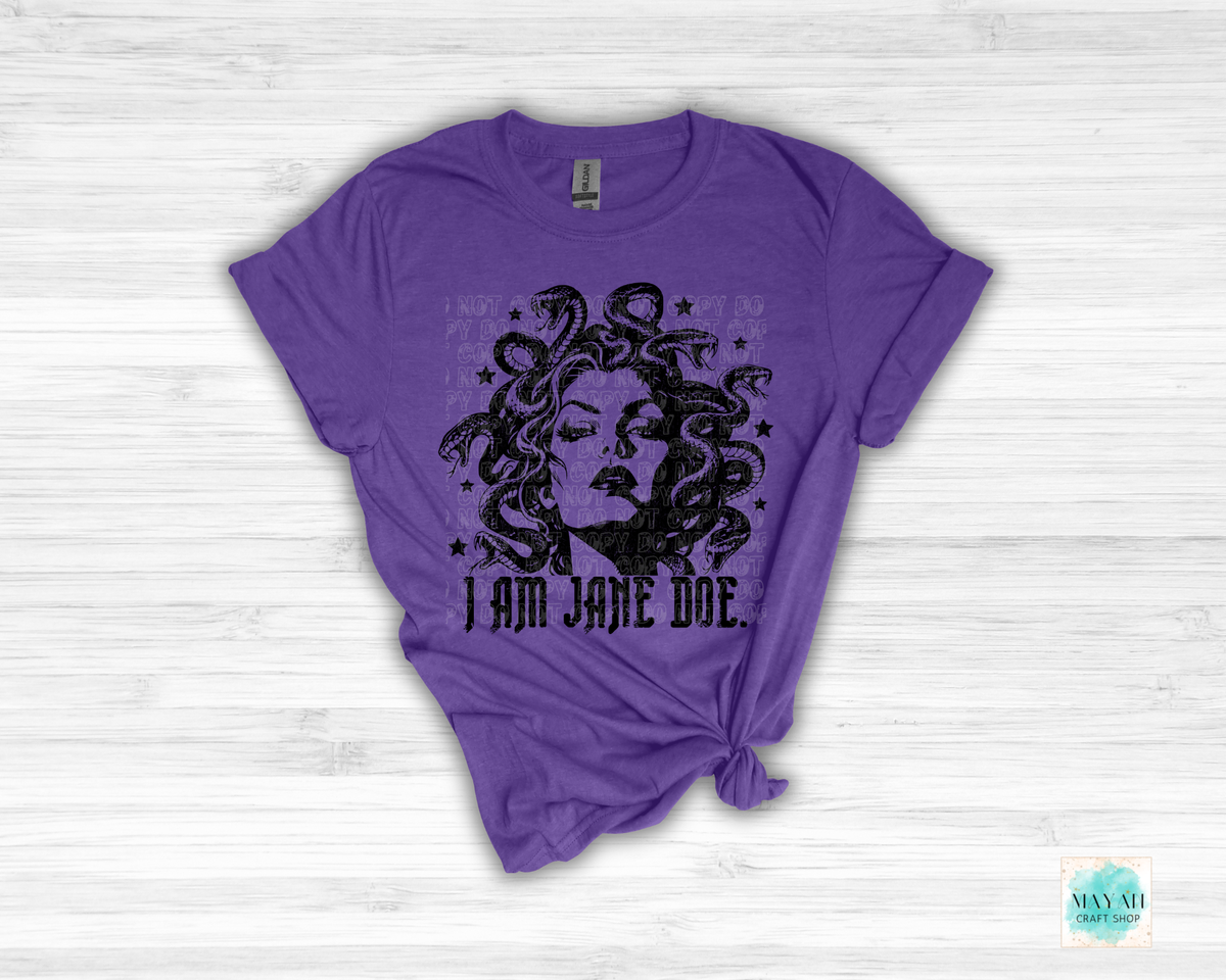 A heather purple short-sleeve t-shirt with a distressed black graphic of Medusa's face surrounded by snakes, with the text 'I AM JANE DOE.' printed below.