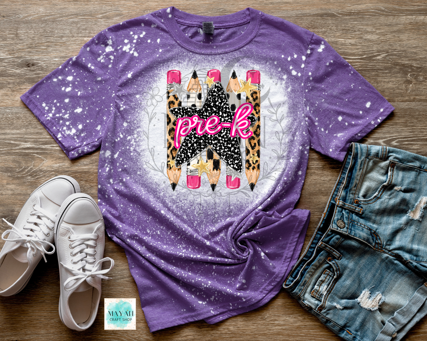 A purple short-sleeve graphic T-shirt with a white bleach-splatter finish, featuring a central design of pencils with leopard print, pink tips, and the word 'pre-k' in pink script. The shirt is styled in a flat lay on a wood background with white sneakers, distressed denim shorts.