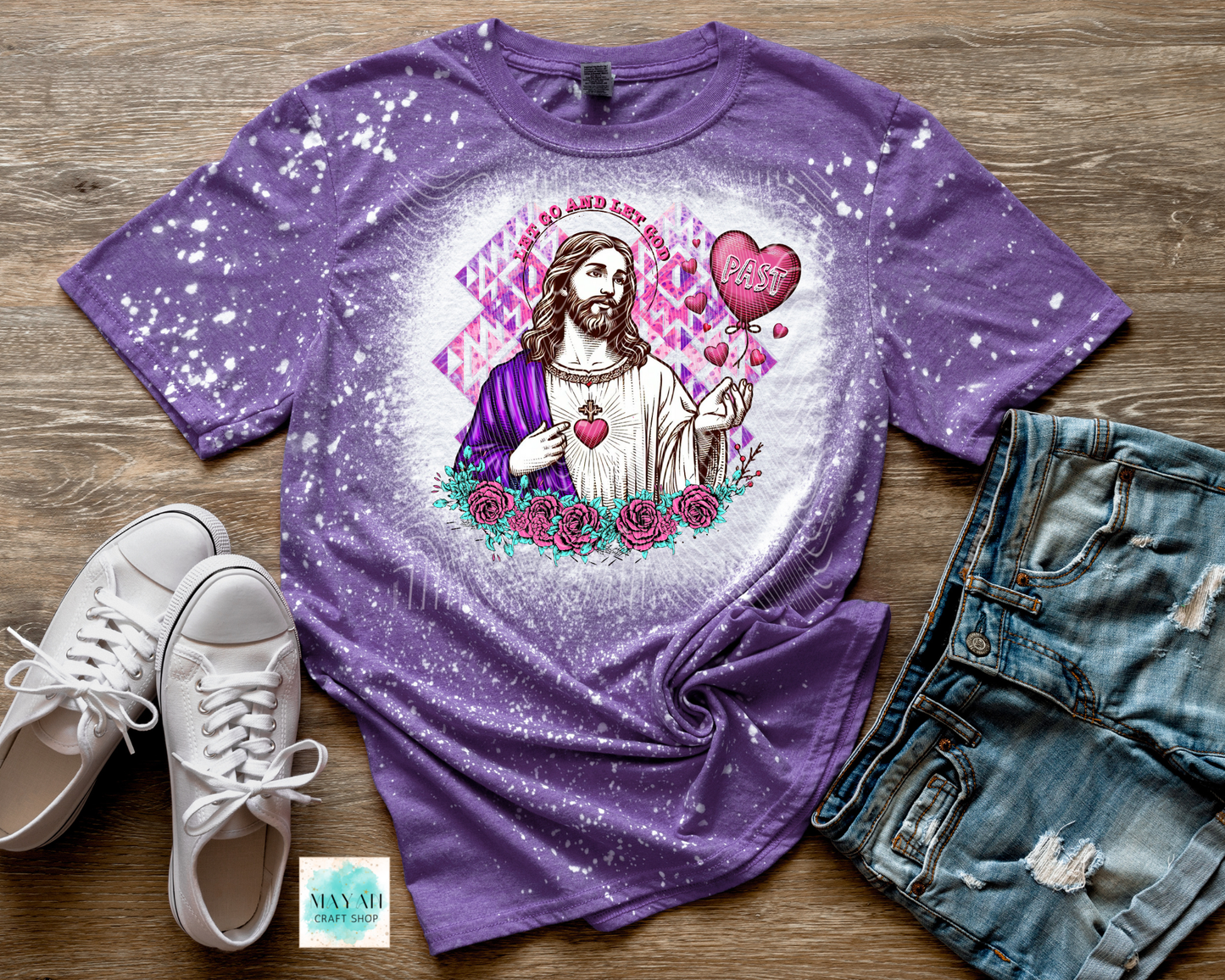 Purple t-shirt with a graphic design of a religious figure, hearts, and flowers on a wooden surface with white sneakers and denim shorts.