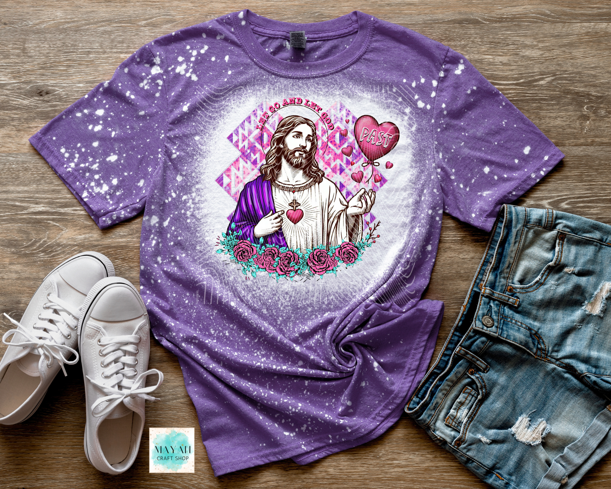 Purple t-shirt with a graphic design of a religious figure, hearts, and flowers on a wooden surface with white sneakers and denim shorts.