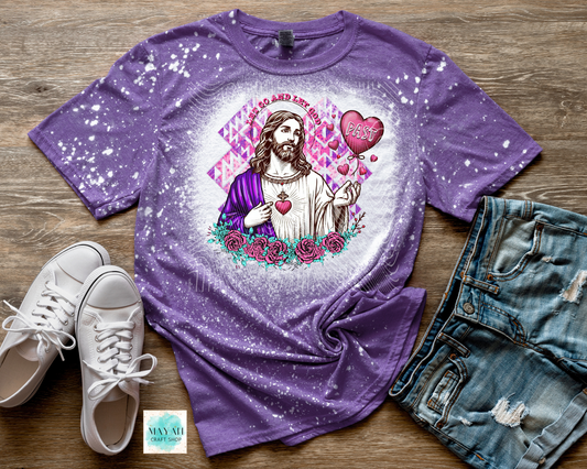 Purple t-shirt with a graphic design of a religious figure, hearts, and flowers on a wooden surface with white sneakers and denim shorts.
