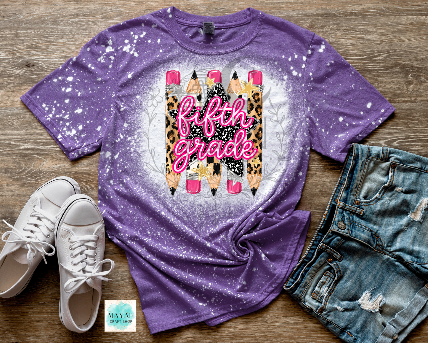 A purple short-sleeve t-shirt with a white splatter bleach effect and a central graphic of pencils with leopard print, pink lettering reading 'fifth grade', and a white circular glow. The shirt is styled in a flat lay on a wood background with white sneakers.
