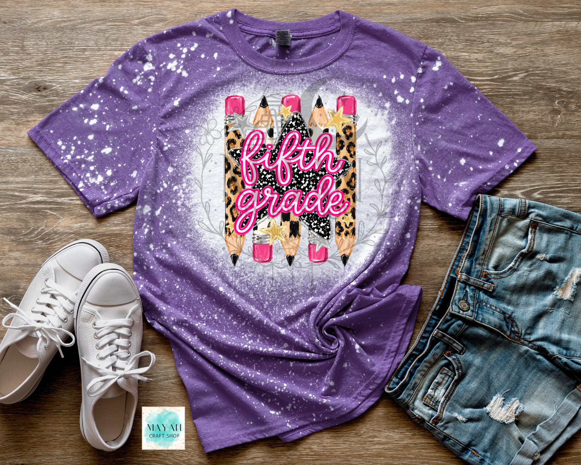 A purple short-sleeve t-shirt with a white splatter bleach effect and a central graphic of pencils with leopard print, pink lettering reading 'fifth grade', and a white circular glow. The shirt is styled in a flat lay on a wood background with white sneakers.