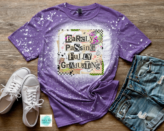 Purple graphic t-shirt with 'Barely Passing Fully Graduating' text, white sneakers, and denim shorts on a wooden surface.