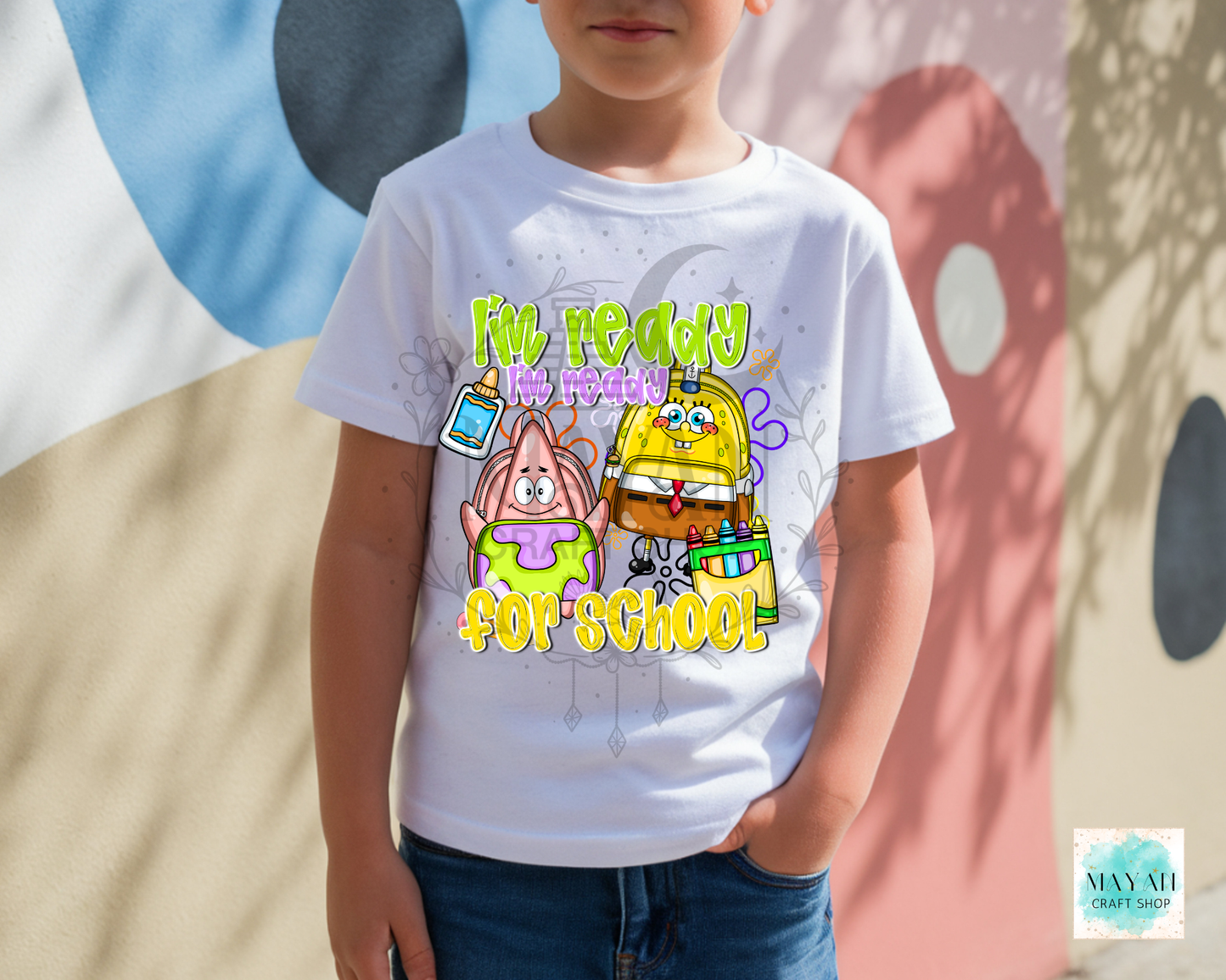 A child wearing a white graphic t-shirt featuring cartoon saying READY FOR SCHOOL' in yellow and green. The child is standing outdoors against a wall with painted shapes.