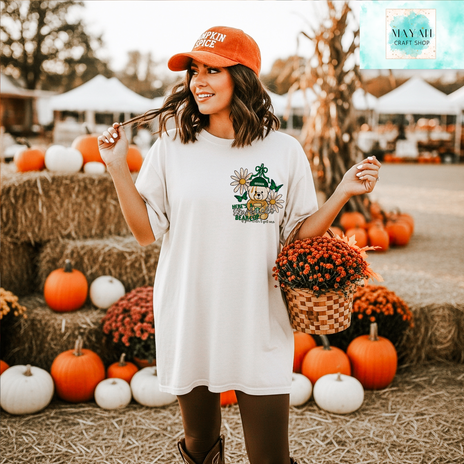 Woman in a white shirt with a graphic design, orange cap, and basket of flowers at a pumpkin patch.