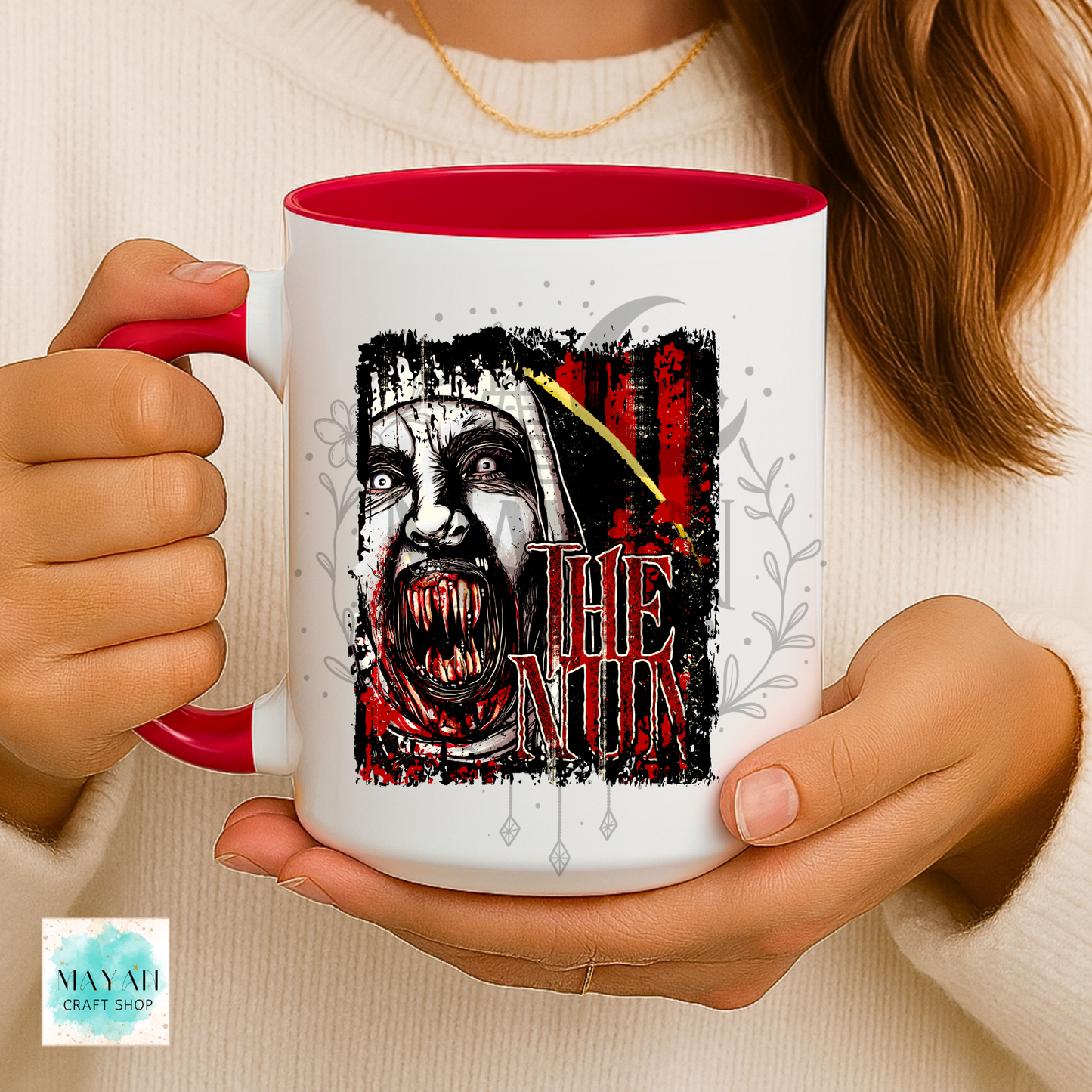 A person holding a white ceramic mug with a red interior and handle. The mug features a graphic of a creepy nun with a knife and a text.