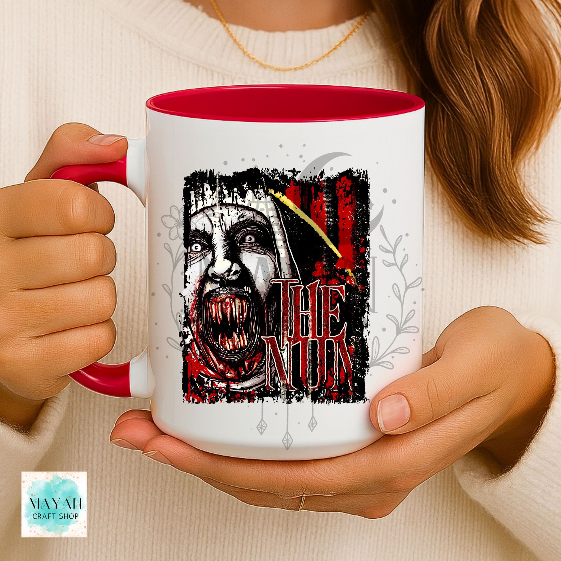 A person holding a white ceramic mug with a red interior and handle. The mug features a graphic of a creepy nun with a knife and a text.