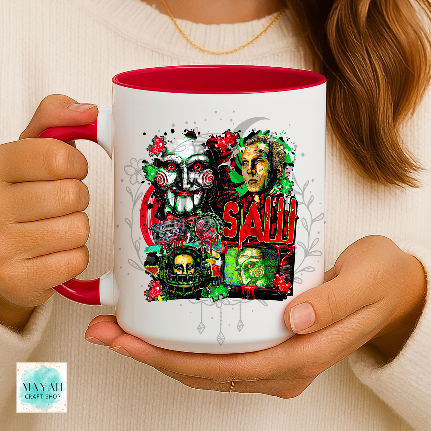 A person holding a white ceramic mug with a red interior and handle. The mug features a colorful graphic collage of horror movie characters, including a clown mask, a green-eyed monster, a skull, and the a text in red.