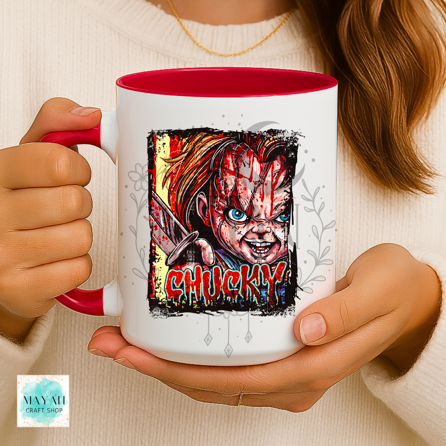 A person holds a white ceramic mug with a red interior and handle. The mug features a graphic illustration of a doll with a word in red dripping letters.