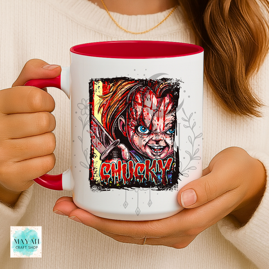 A person holds a white ceramic mug with a red interior and handle. The mug features a graphic illustration of a doll with a word in red dripping letters.