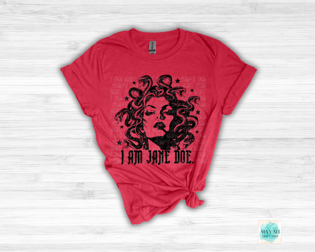 A heather red short-sleeve t-shirt with a black graphic of a woman's face surrounded by snakes and the text 'I AM JANE DOE.' printed on the front, styled flat on a white wood background.