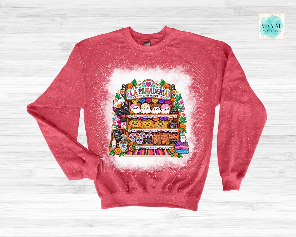 A heathered coral-pink crewneck sweatshirt featuring a colorful graphic of a Halloween-themed bakery stand labeled 'La Panadería'. The sweatshirt is displayed flat on a light gray wood-plank background with a white paint-splatter effect around the central graphic.