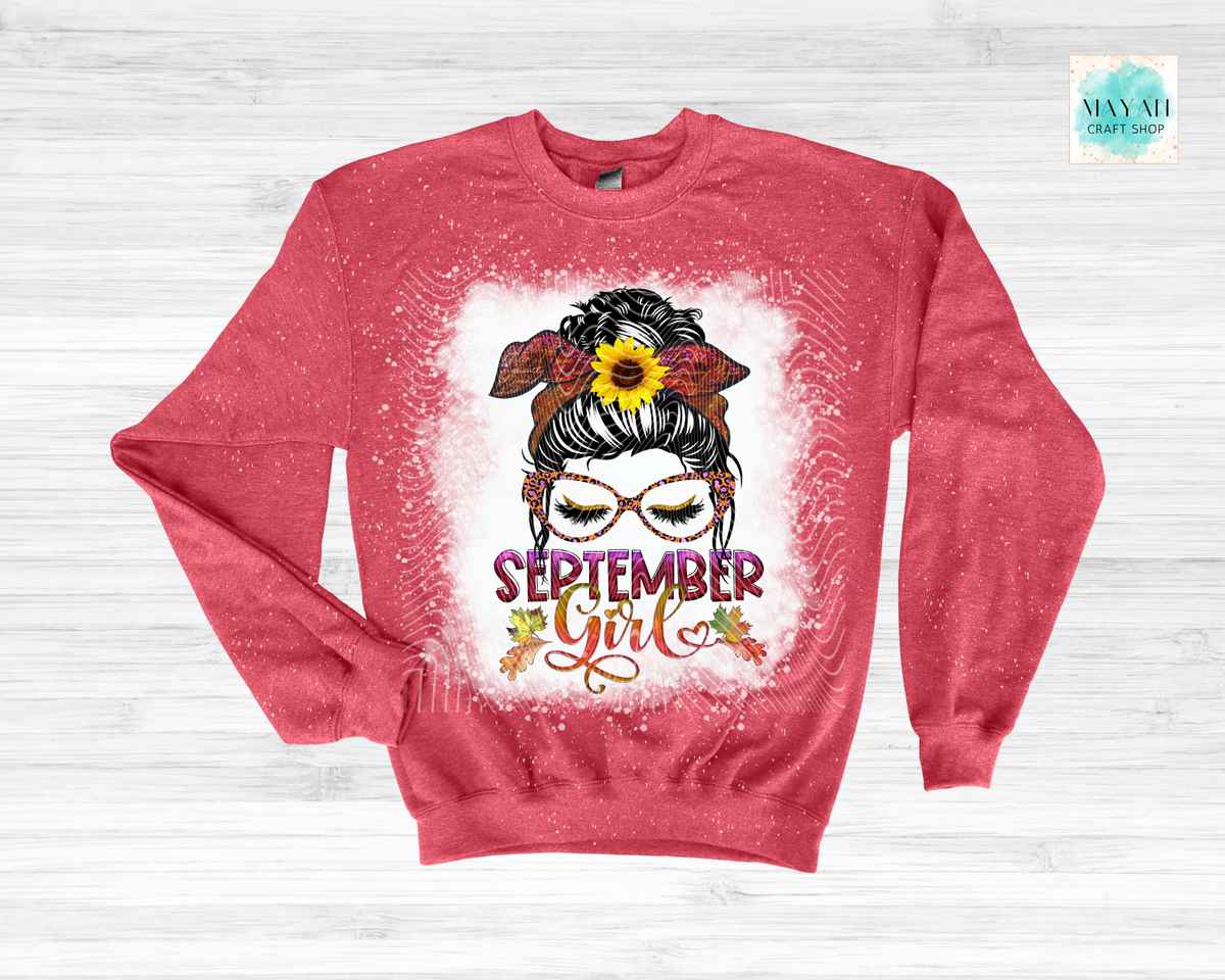 A heathered red crewneck sweatshirt with a distressed white paint-splatter graphic on the front. The graphic features a stylized woman with glasses, a sunflower in her hair, and the text 'SEPTEMBER girl' in purple and orange script.