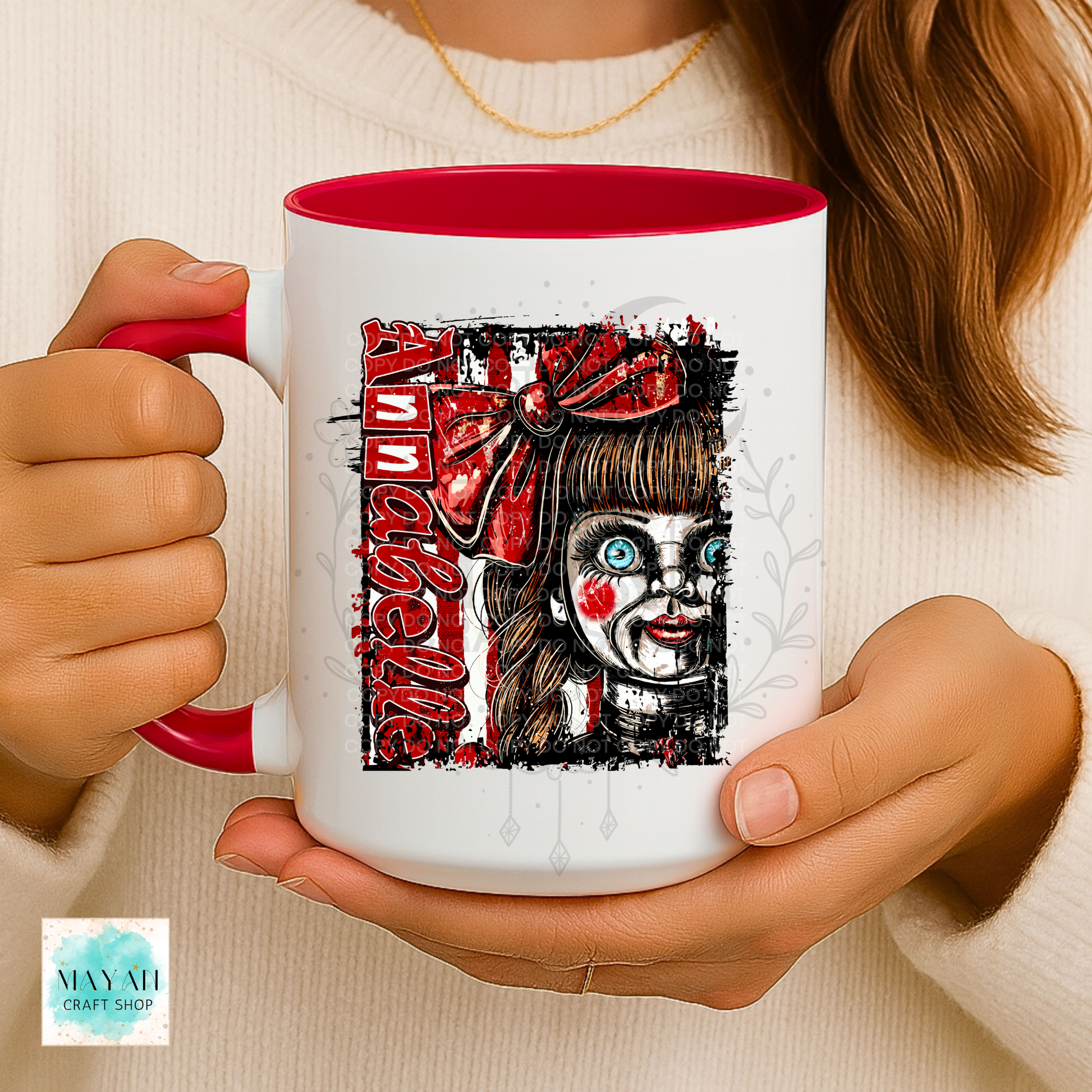 A person holds a white ceramic mug with a red interior and handle. The mug features a graphic illustration of a creepy doll-like face with red accents and a word in red block letters.