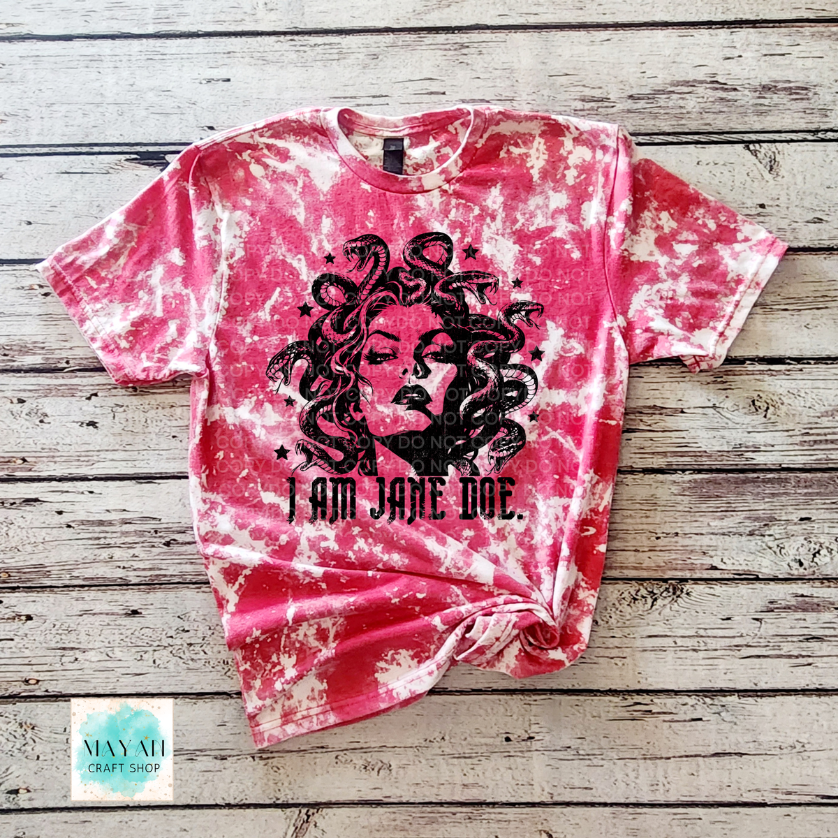 A heather red bleached t-shirt with a black graphic of Medusa and the text 'I AM JANE DOE' printed on the front, laid flat on a rustic wooden background.