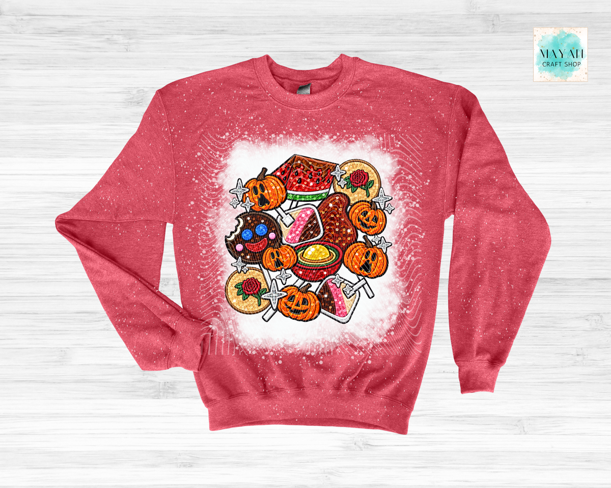 A heathered red crewneck sweatshirt featuring a colorful Halloween-themed graphic print of jack-o'-lanterns, candy corn, a pumpkin pie, and donuts on a white splattered background, laid flat on a light gray wood-textured surface.