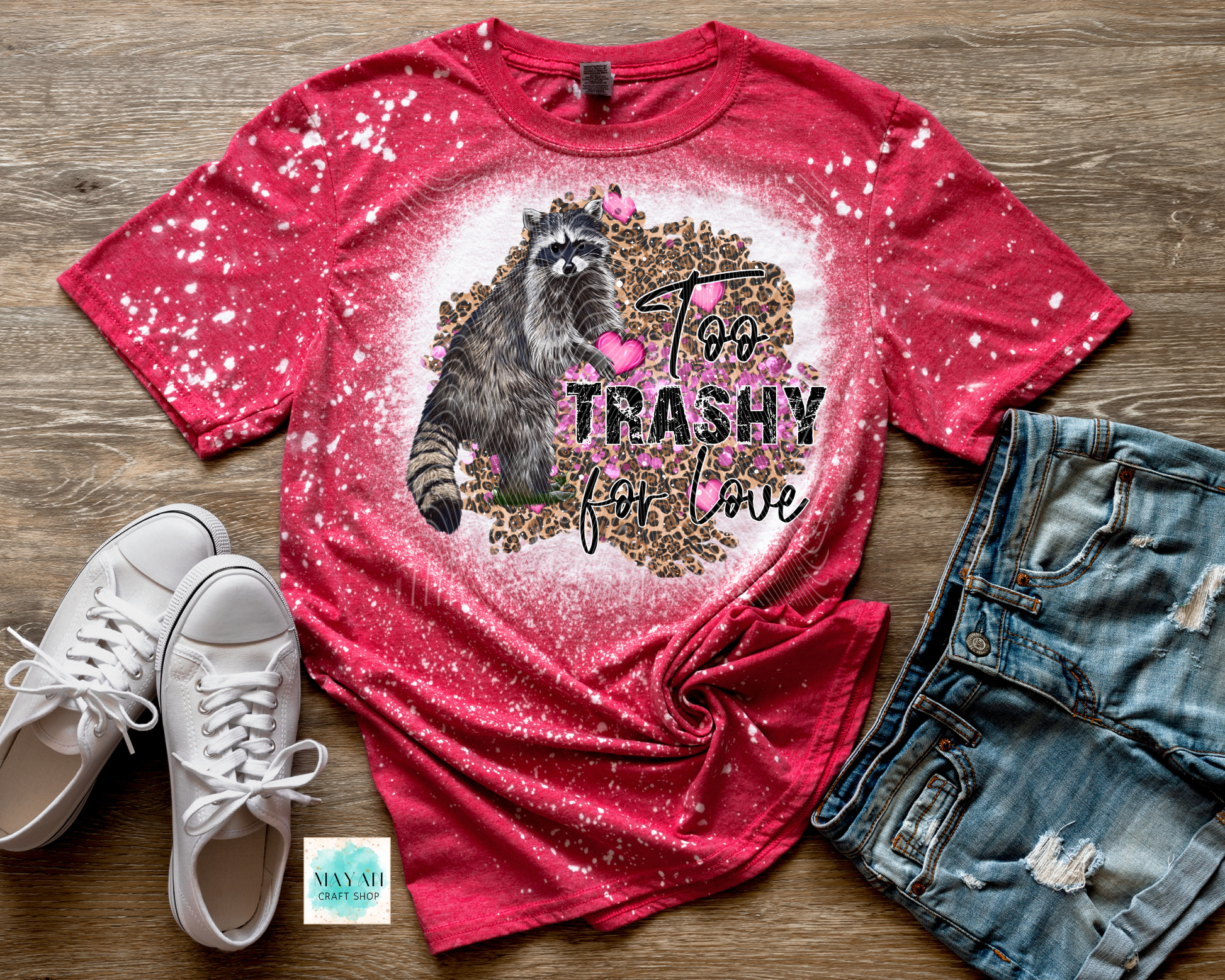 Red t-shirt with a raccoon graphic and text, paired with white sneakers and denim shorts on a wooden surface.