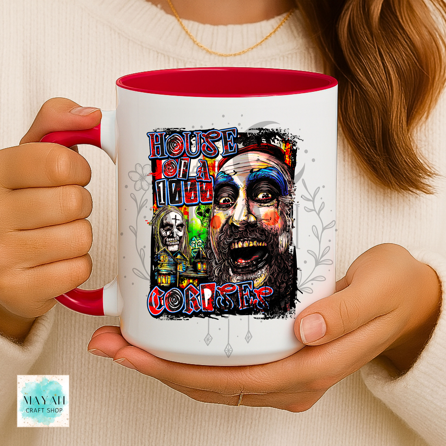 A person holds a white ceramic mug with a red interior and handle. The mug features a colorful graphic print with a text and a stylized illustrated face.