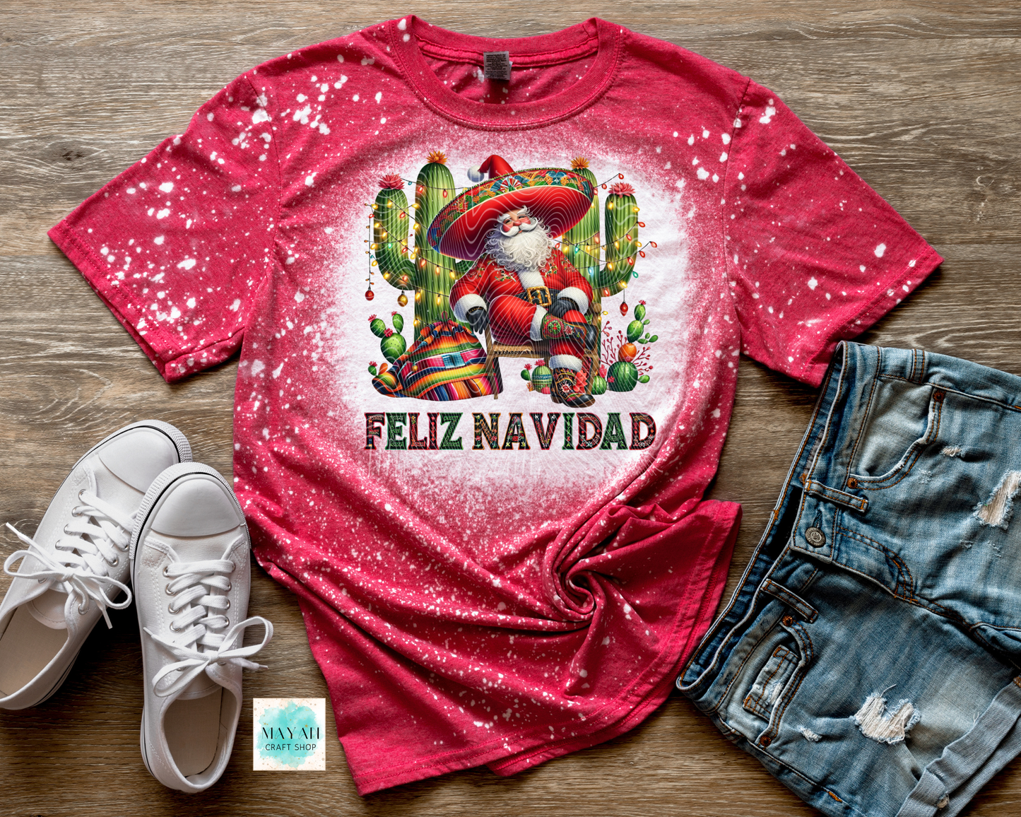 Red t-shirt with Santa Claus and cactus design, 'Feliz Navidad' text, white sneakers, and denim shorts on a wooden surface.