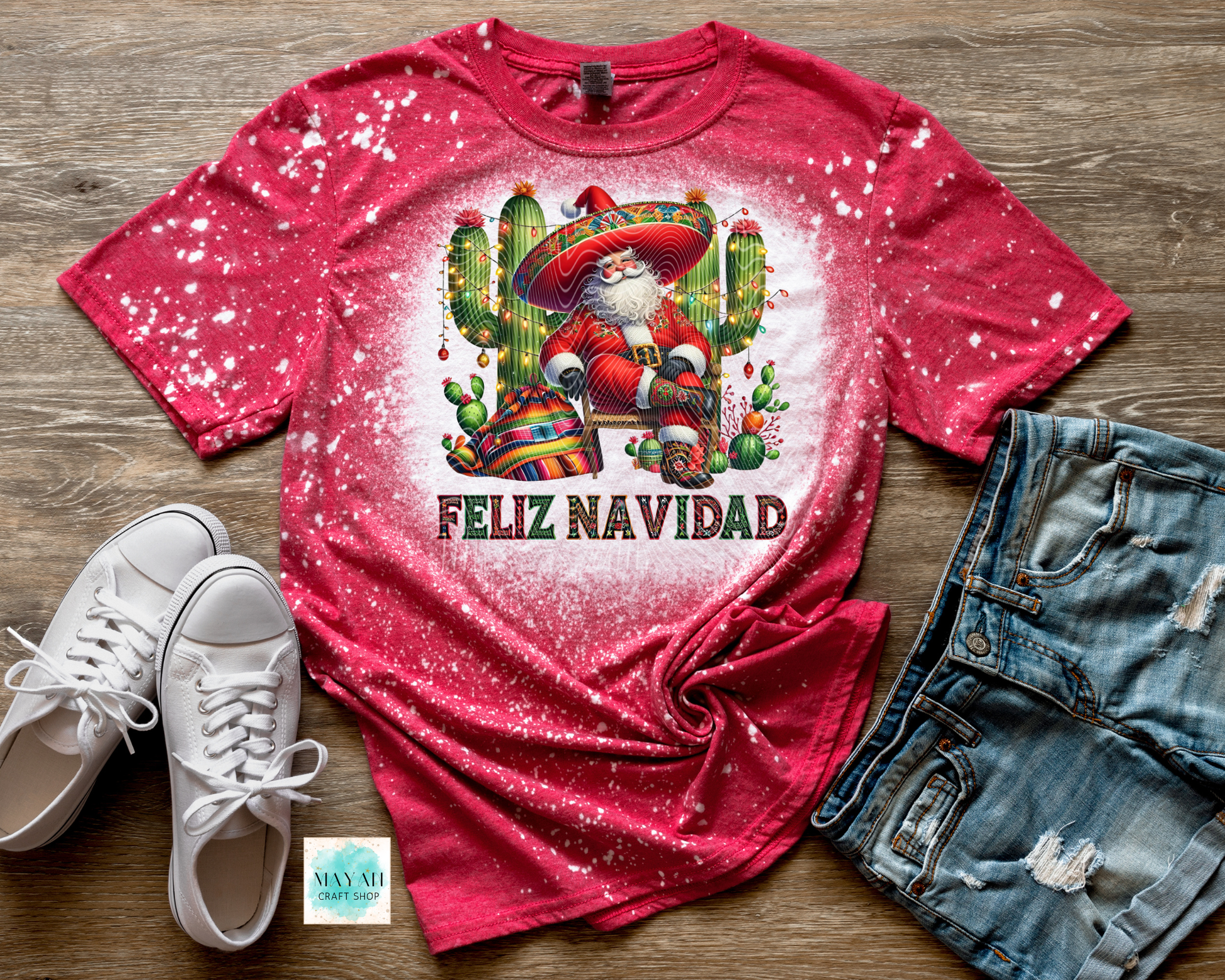 Red t-shirt with Santa Claus and cactus design, 'Feliz Navidad' text, white sneakers, and denim shorts on a wooden surface.