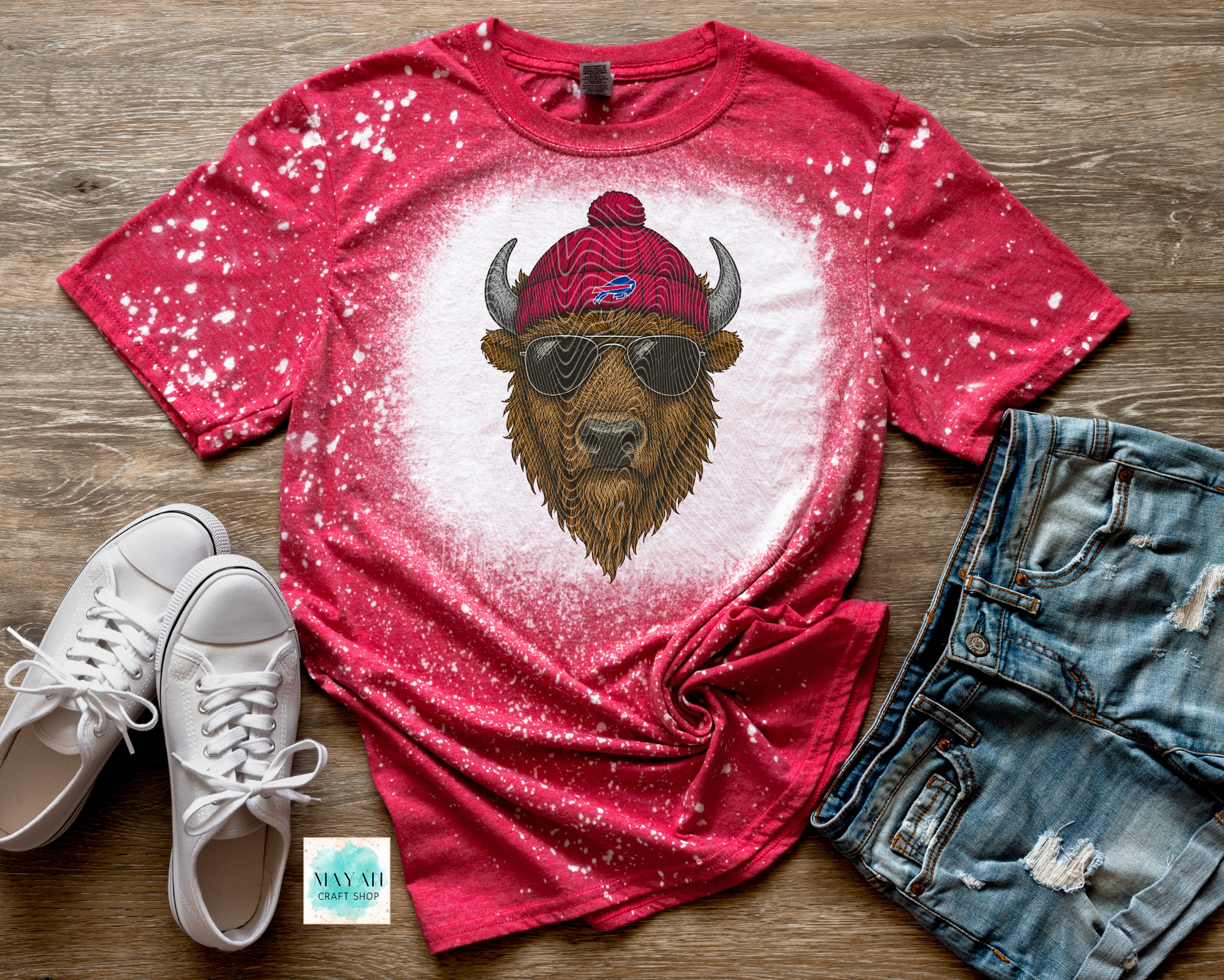 Red t-shirt with a bear graphic wearing sunglasses and a beanie, paired with white sneakers and denim shorts on a wooden surface.