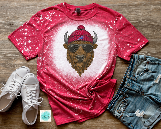 Red t-shirt with a bear graphic wearing sunglasses and a beanie, paired with white sneakers and denim shorts on a wooden surface.