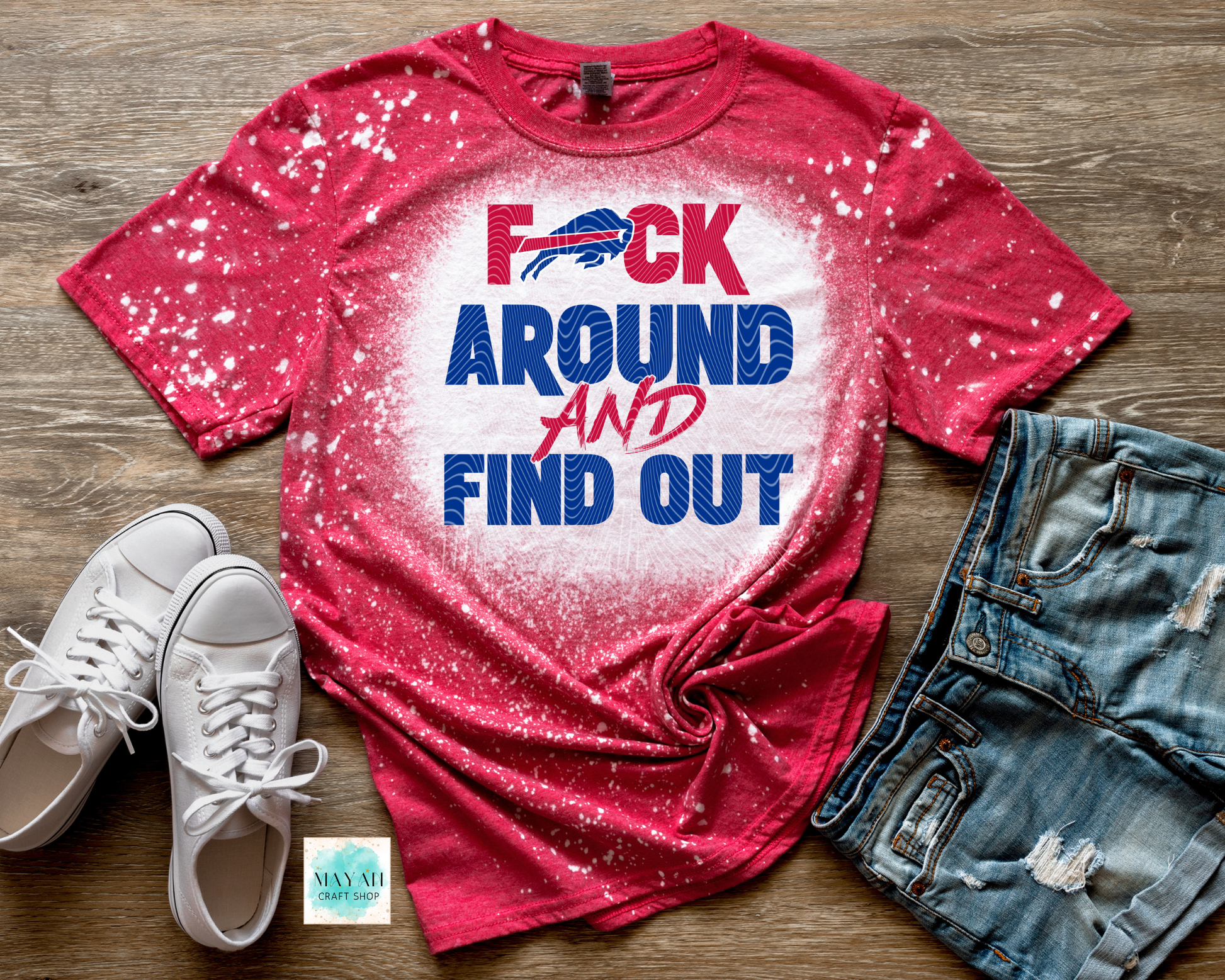 Red t-shirt with graphic design and text on a wooden surface with white sneakers and denim shorts.