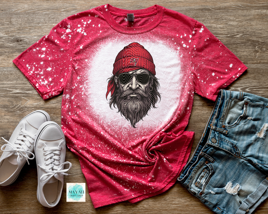 Red t-shirt with graphic design of a bearded man wearing a cap, paired with white sneakers and denim shorts on a wooden surface.