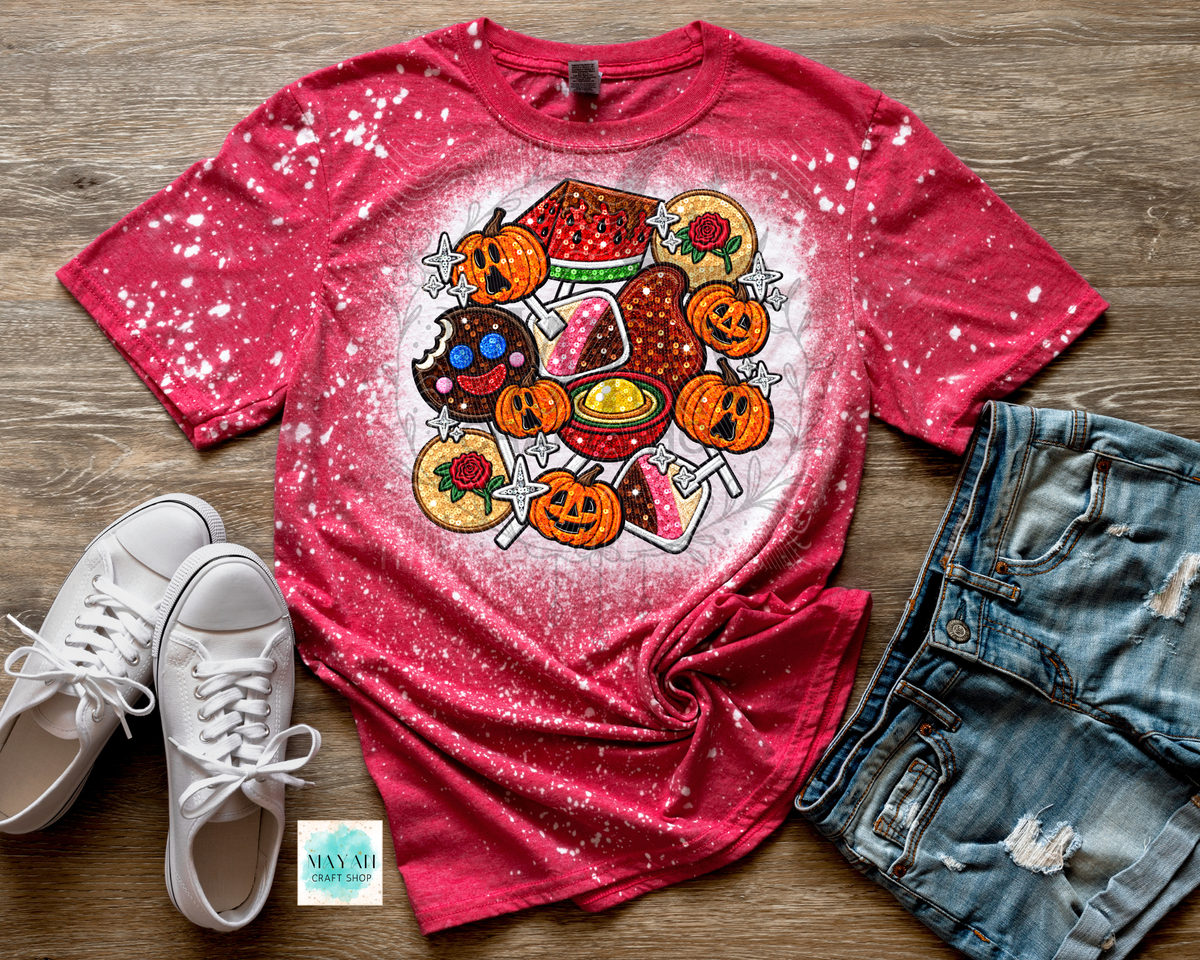 A styled flat-lay of a short-sleeve graphic T-shirt on a wood floor, paired with white sneakers and distressed denim shorts. The shirt is bright pink with a white glittery splatter effect and features a large colorful Halloween-themed food graphic on the front, including pumpkins, candy corn, donuts, and treats.