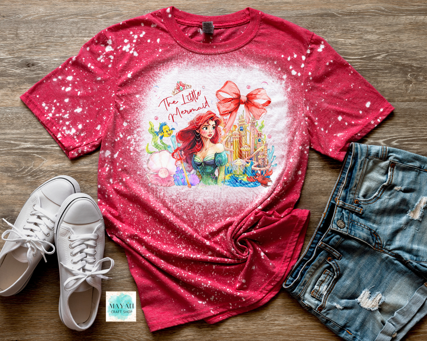 Red t-shirt with a mermaid design on a wooden surface with white sneakers and denim shorts.