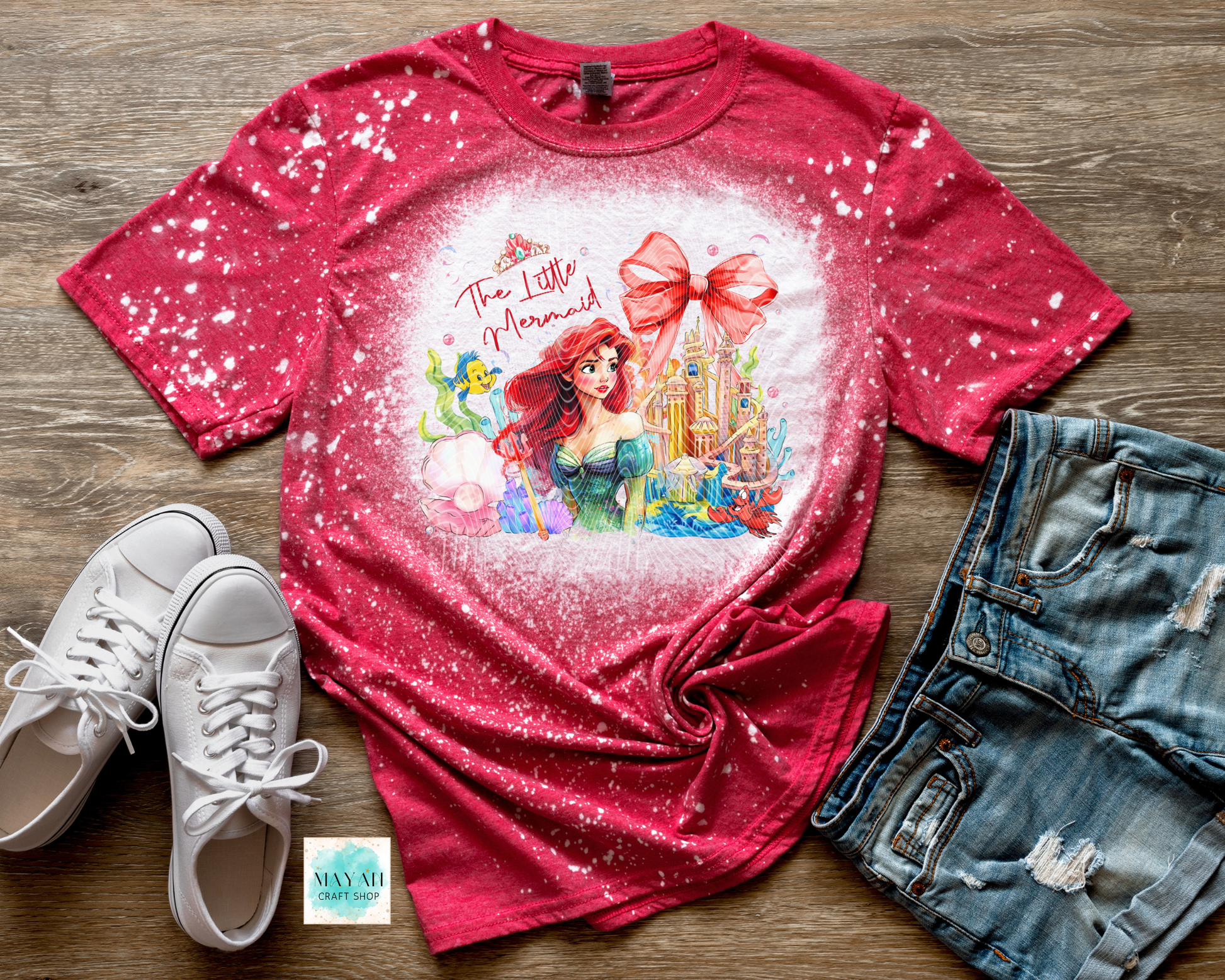 Red t-shirt with a mermaid design on a wooden surface with white sneakers and denim shorts.