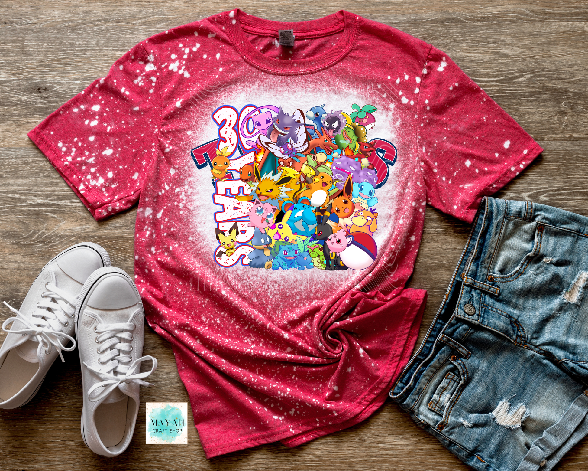 Red t-shirt with colorful graphic design on a wooden surface with white sneakers and denim shorts.