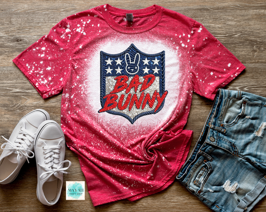 Red t-shirt with 'Bad Bunny' logo on a wooden surface with white sneakers and denim shorts.