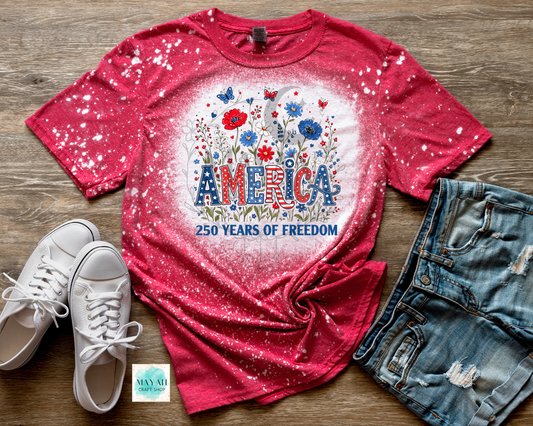 Red t-shirt with 'America 250 Years of Freedom' design on a wooden surface with white sneakers and denim shorts.