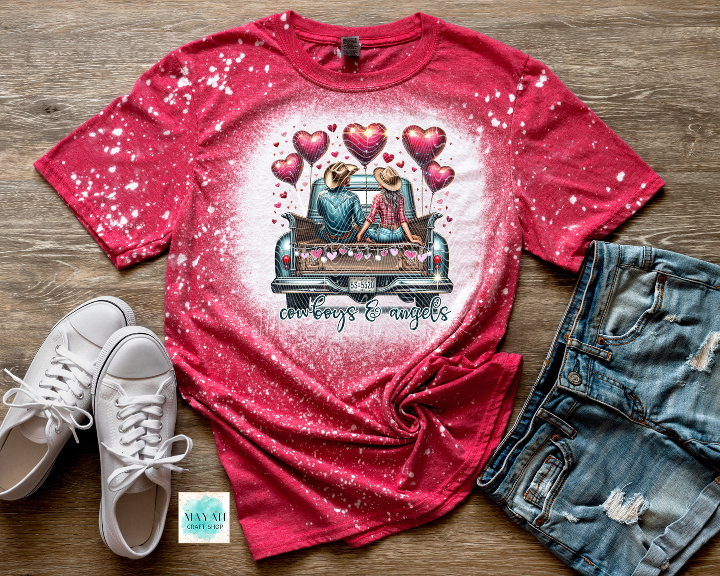 Red t-shirt with graphic design of a couple in a truck surrounded by hearts, paired with white sneakers and denim shorts on a wooden surface.