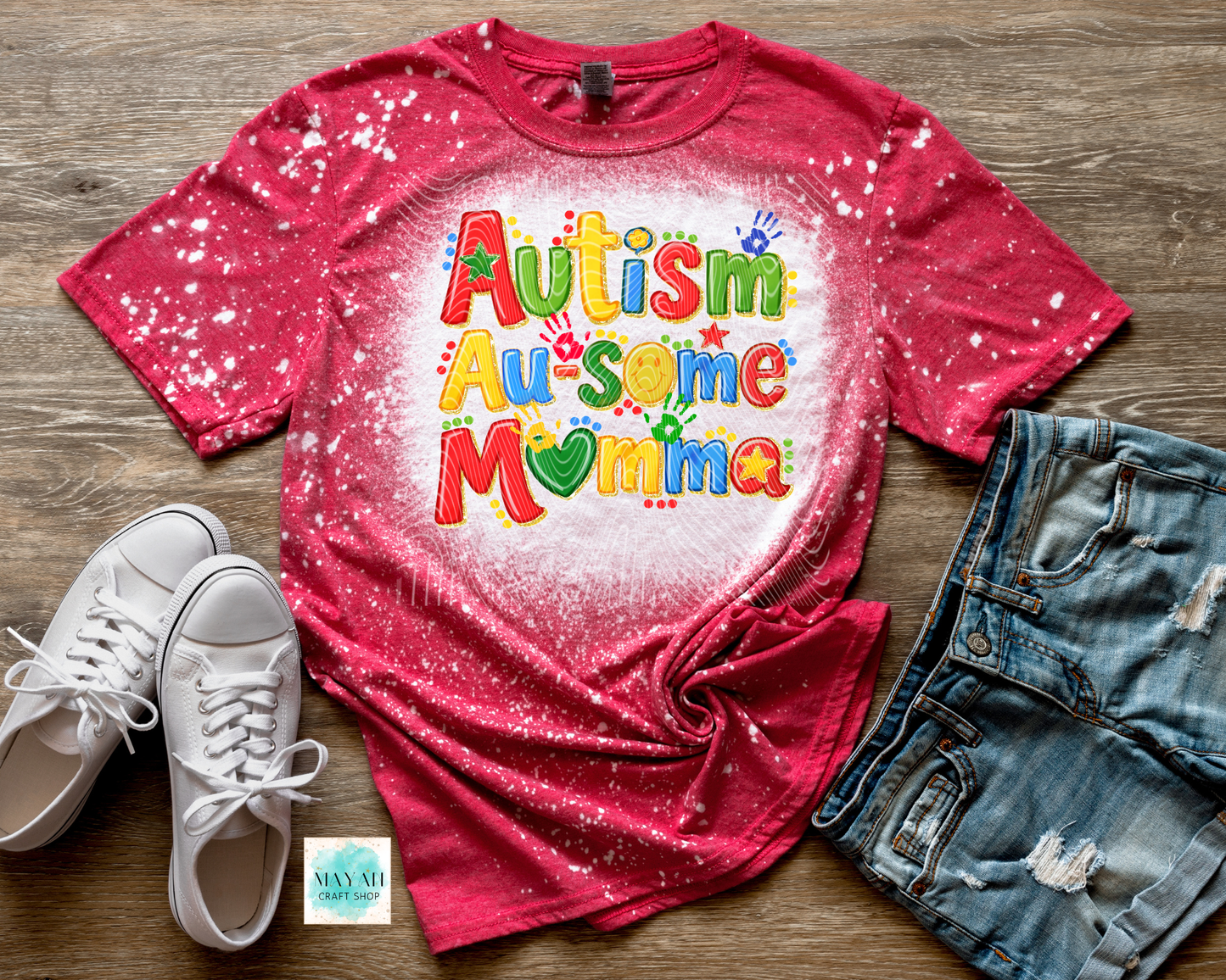 Red t-shirt with 'Autism Awesome Mama' text on a wooden surface with shoes and shorts.