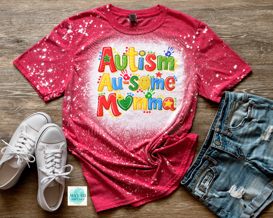Red t-shirt with 'Autism Awesome Mama' text on a wooden surface with shoes and shorts.