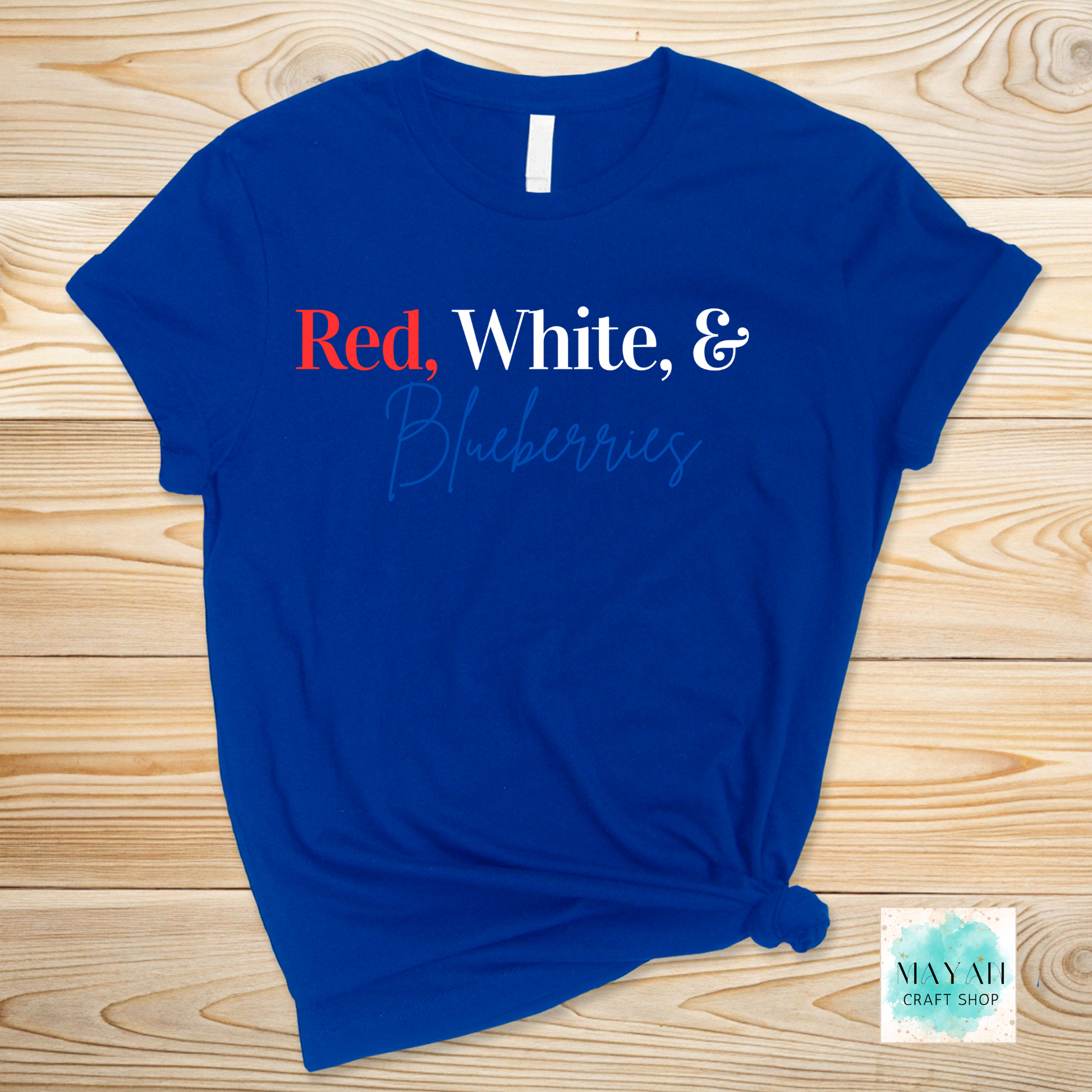 Blue t-shirt with 'Red, White, & Blueberries' text on a wooden surface, featuring the MayNH Craft Shop logo.