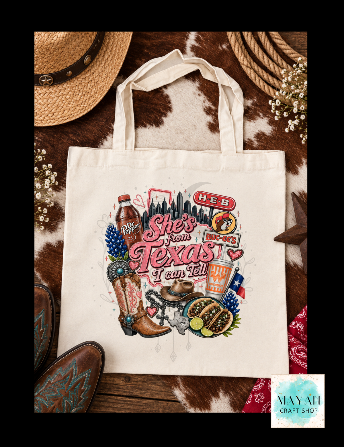 A cream canvas tote bag with a colorful 'She's from Texas' graphic, styled in a flat-lay on a cowhide background with boots, a hat, rope, and flowers.