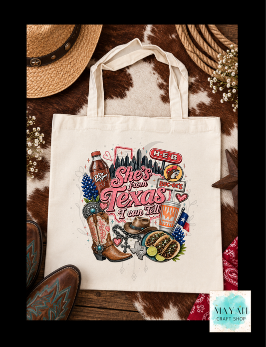 A cream canvas tote bag with a colorful 'She's from Texas' graphic, styled in a flat-lay on a cowhide background with boots, a hat, rope, and flowers.