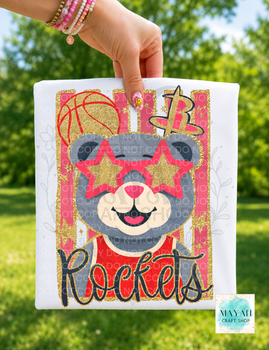 A hand holds a white shirt featuring a cartoon bear wearing pink star glasses and a red shirt, with a word in script below. The background is an outdoor grassy area with blurred green trees.