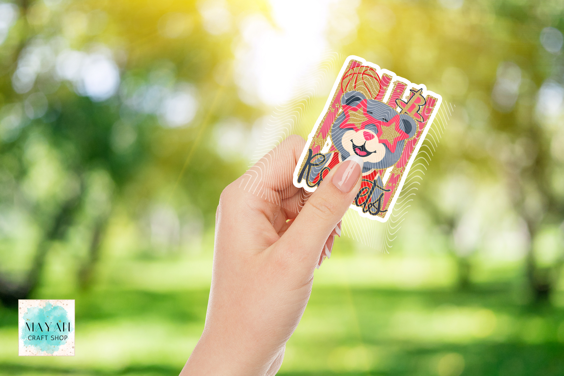 A hand holds a die-cut sticker featuring a colorful cartoon bear wearing a red crown, set against a bright, sunlit green park background with a soft bokeh effect. 