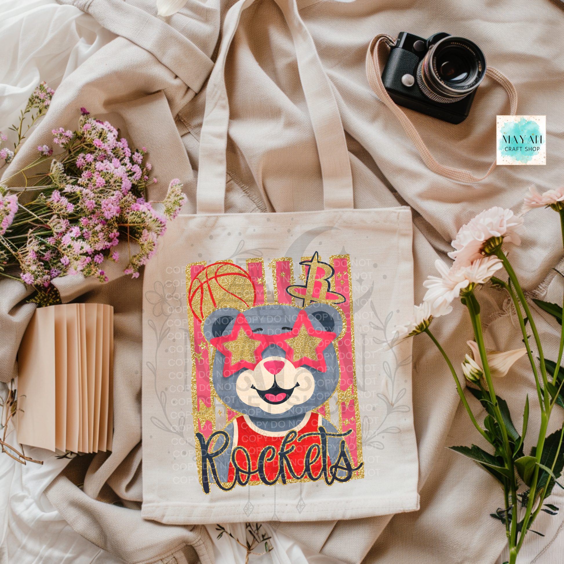 A canvas tote bag with a colorful embroidered bear face and a word in script, styled on beige fabric with flowers, a book, and a camera.