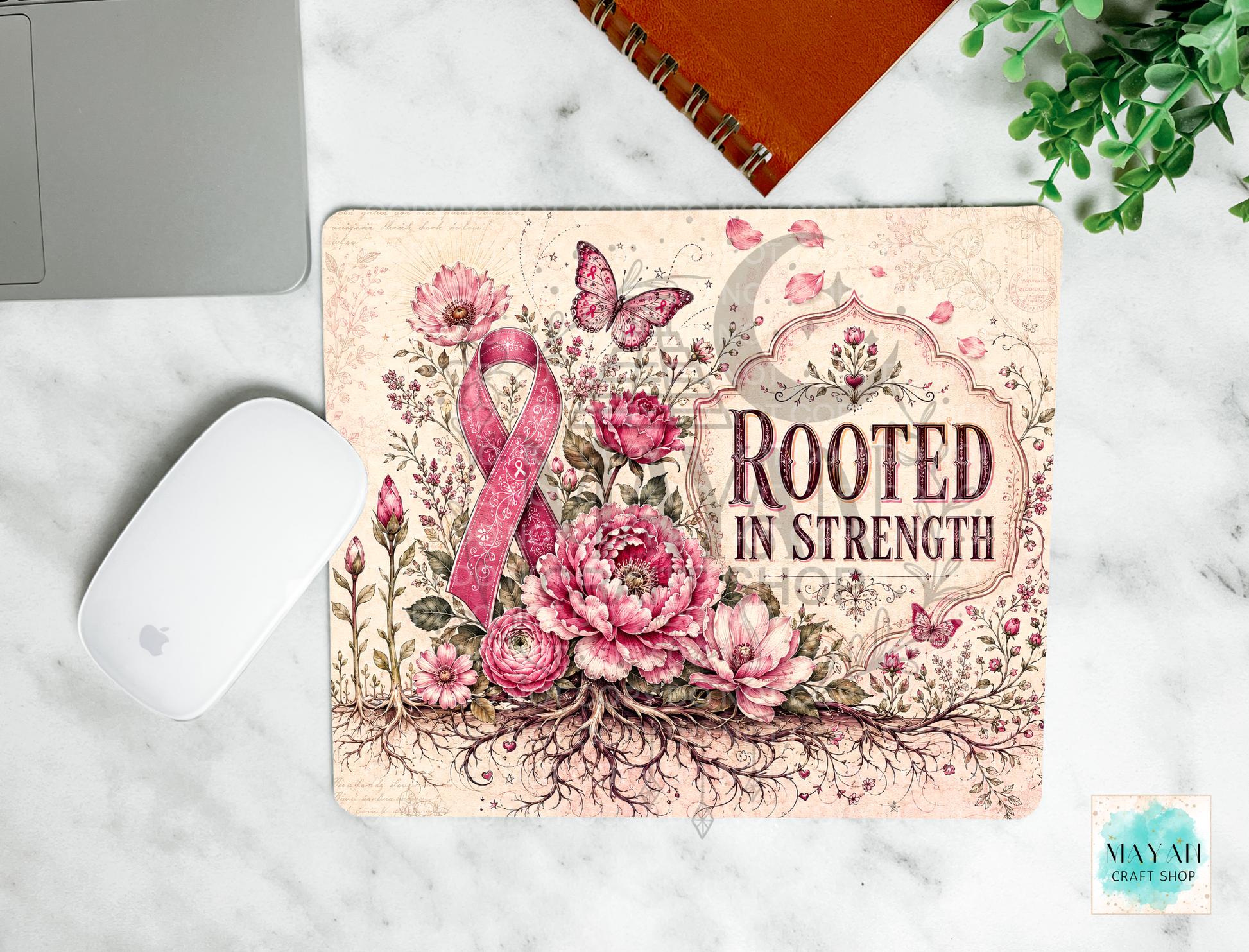 A rectangular mouse pad with a floral and ribbon design sits on a white marble surface. The mat features pink and beige flowers, butterflies, a pink awareness ribbon, and the words 'ROOTED IN STRENGTH.