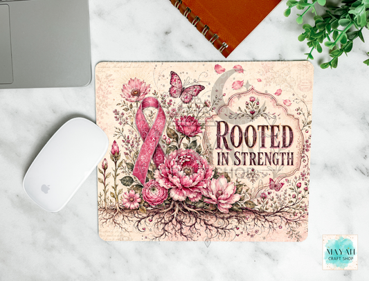 A rectangular mouse pad with a floral and ribbon design sits on a white marble surface. The mat features pink and beige flowers, butterflies, a pink awareness ribbon, and the words 'ROOTED IN STRENGTH.