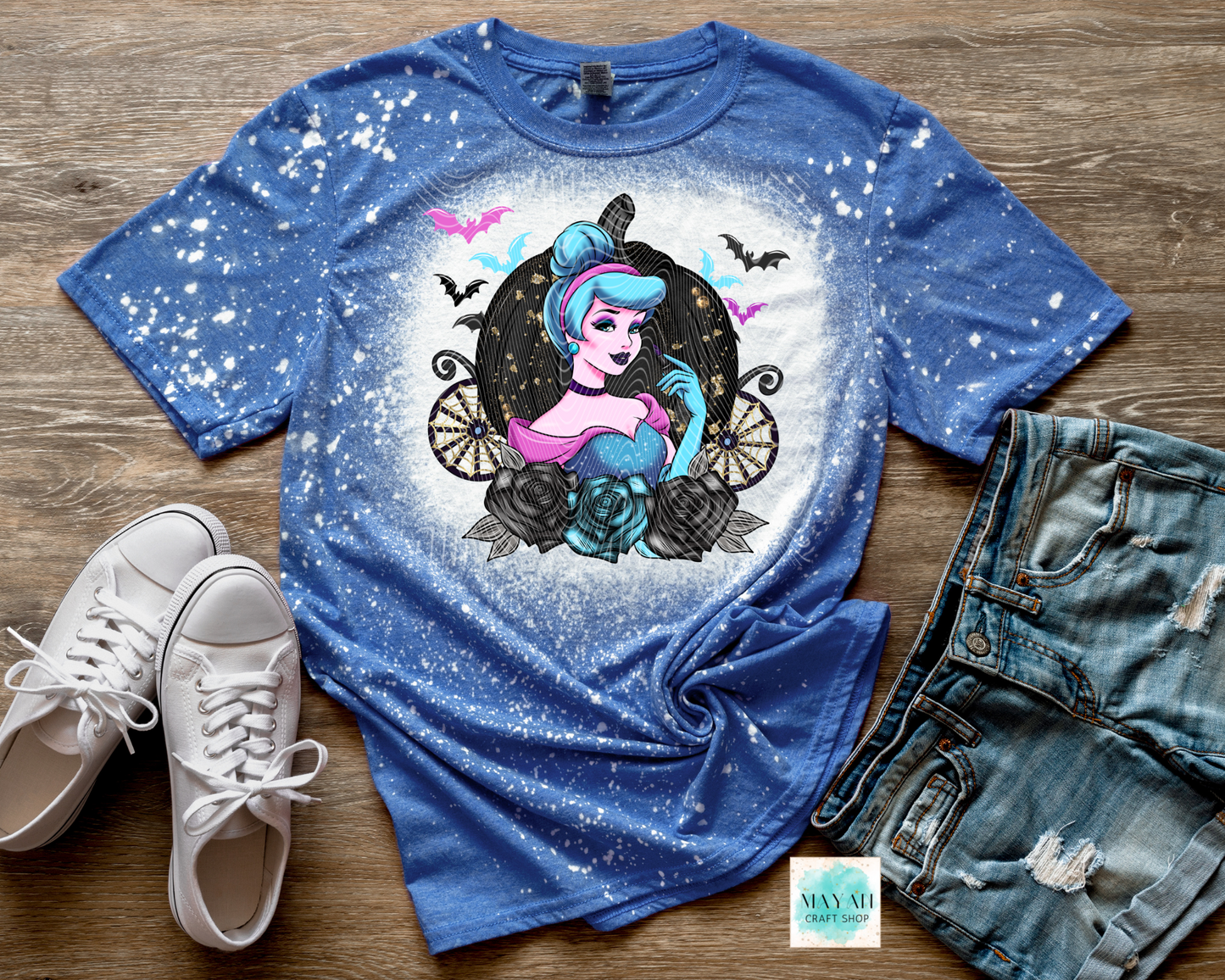 Blue t-shirt with a graphic design of a character in a circular frame, surrounded by bats and flowers, on a wooden surface with white sneakers and denim shorts.