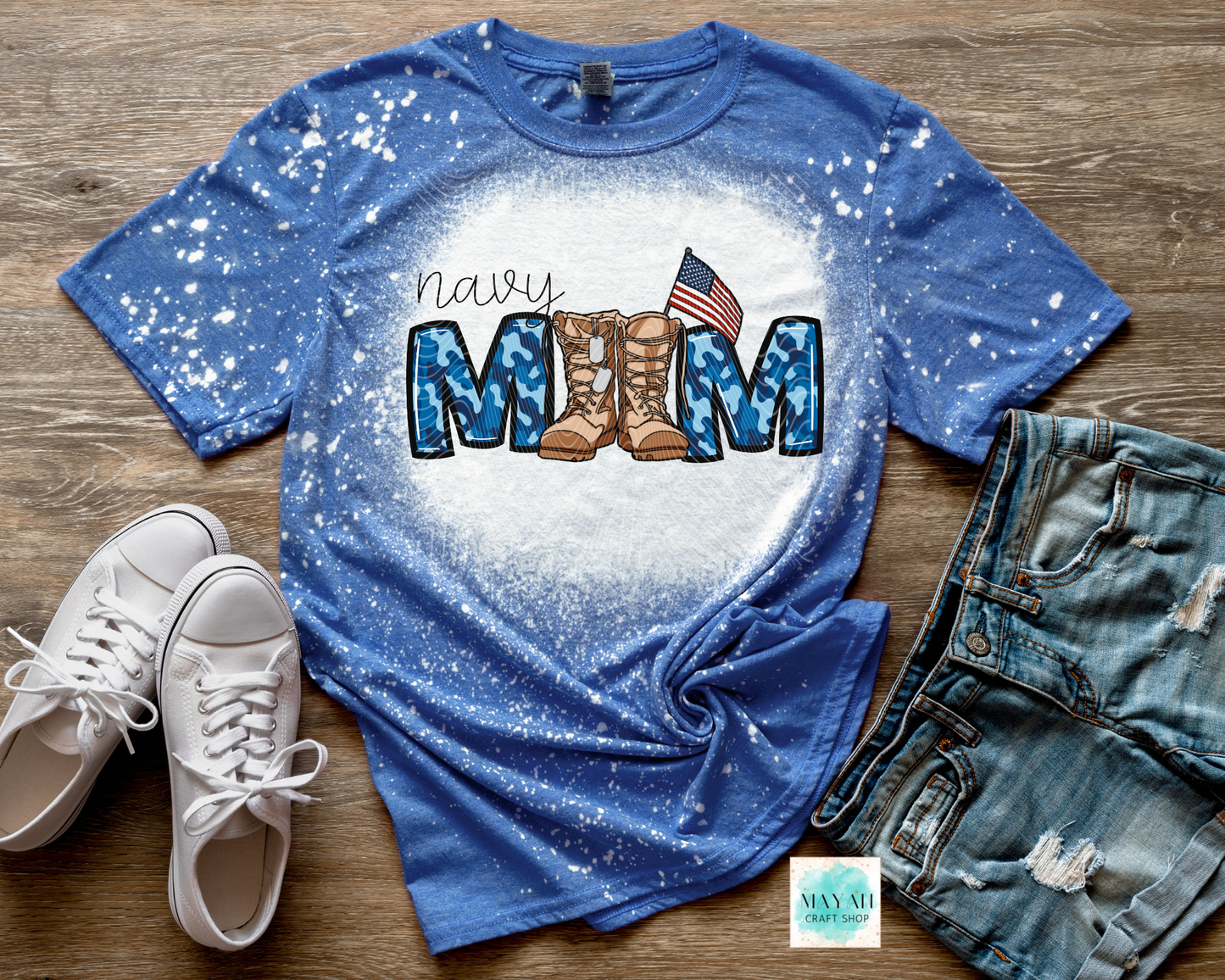 Blue t-shirt with military mom design featuring boots and an American flag, displayed with white sneakers and denim shorts on a wooden surface.